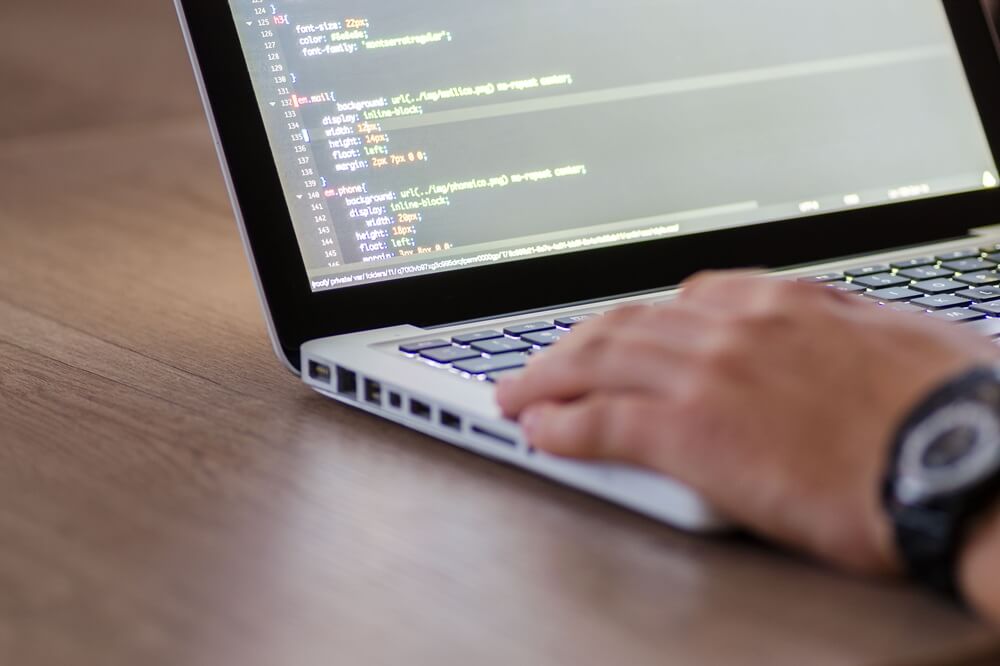 A man on a laptop, his hand is on the bottom right, laptop has code on top half of the screen